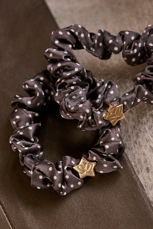 Two dark brown byEloise silk scrunchies with white polka dots and gold star charms, displayed on a textured brown and beige surface.